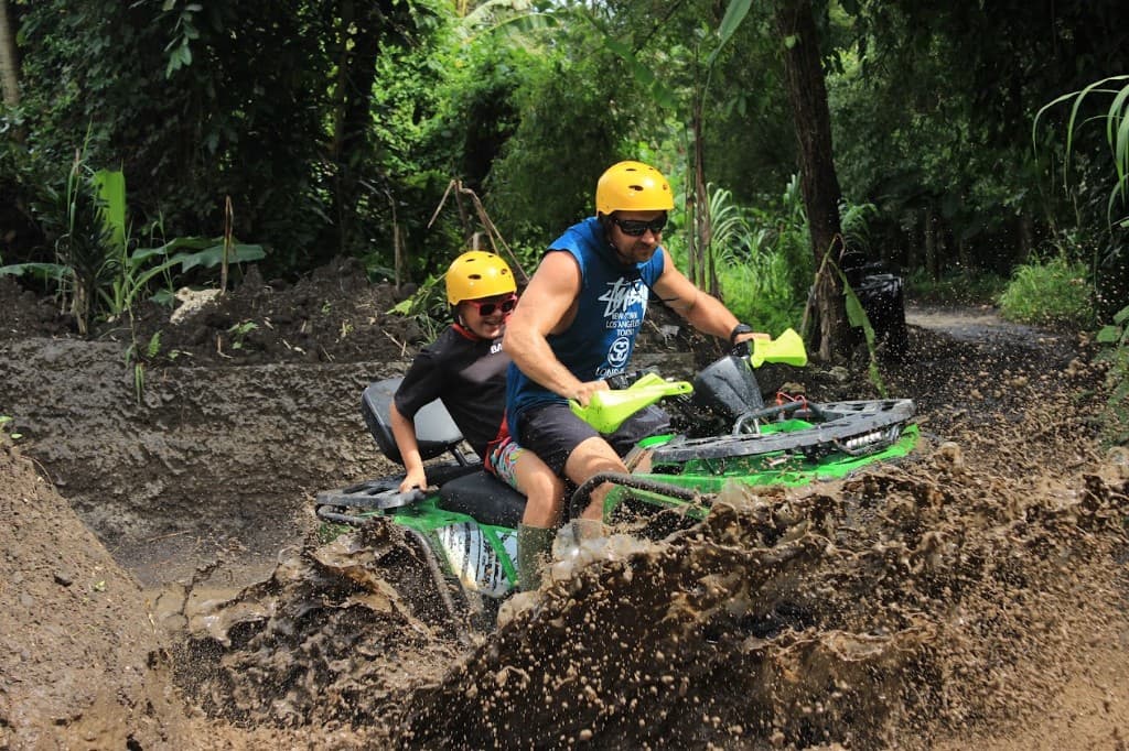 Parent driving a tandem ATV with a child behind through jungle mud