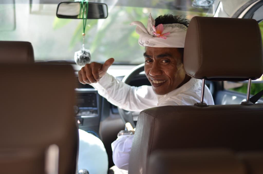 Smiling Balinese guide in a white udeng headcloth giving a thumbs-up, ready to meet guests
